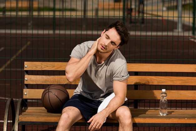 Giocatore di basket seduto su una panca di legno e tenendo un asciugamano e una bottiglia d'acqua al campo da basket