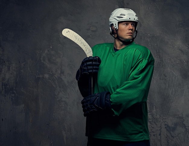 Giocatore di hockey che indossa indumenti protettivi verdi e casco bianco in piedi con la mazza da hockey. Isolato su sfondo grigio.