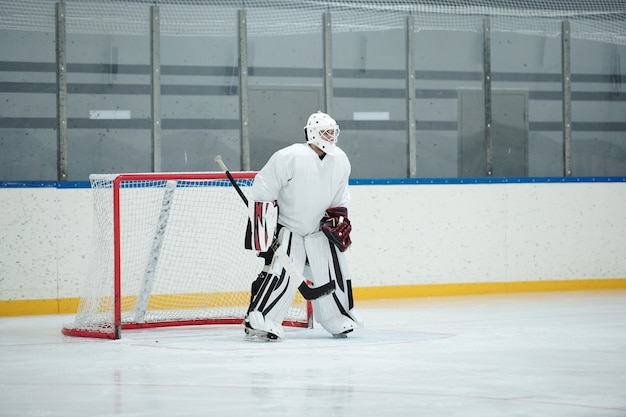 Giocatore di hockey in divisa sportiva bianca, casco protettivo, guanti e pattini che tengono il bastone mentre si trova in piedi sulla pista davanti alla rete e in attesa del disco