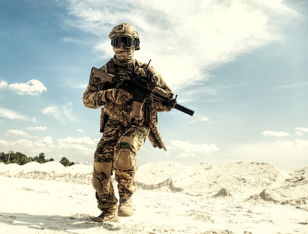 Giocatore di softair in uniforme mimetica protetto con casco e occhiali balistici in esecuzione con replica di fucile di servizio dell'esercito in area sabbiosa durante operazioni militari, collisioni di guerra o simulazione di battaglia