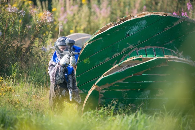 Giocatore di sport di paintball in uniforme protettiva e maschera che giocano con la pistola all'aperto e nascondersi