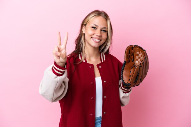 Giocatore donna russa con guanto da baseball isolato su sfondo rosa sorridente e mostrando segno di vittoria