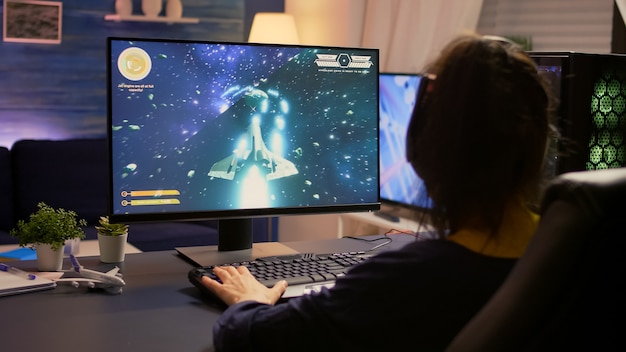 Giocatore seduto su una sedia da gioco e inizia a giocare al videogioco sparatutto spaziale indossando cuffie professionali. Studio moderno dotato di desktop con sistema RGB, tastiera, mouse, utilizzando la tecnologia wirele di rete