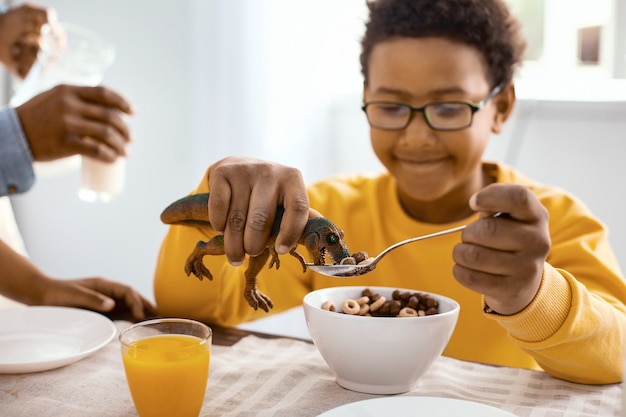 Gioco divertente. Affascinante ragazzo ottimista che gioca con un dinosauro giocattolo che è al centro dell'attenzione e lo alimenta con cereali mentre fa colazione lui stesso