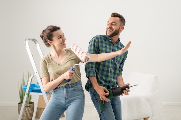 Giocosa giovane donna che canta nel rullo di vernice mentre suo marito usa il trapano come chitarra, si divertono mentre fanno lavori di ristrutturazione