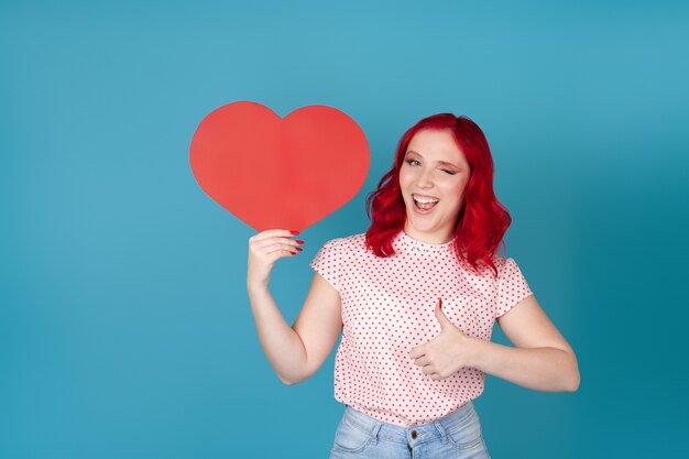 giocosa giovane donna con i capelli rossi che tiene un cuore di carta rossa, ammiccante e dando un pollice in alto