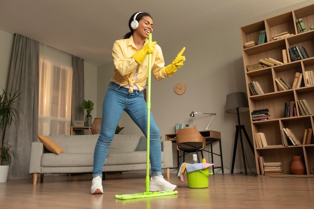 Giocosa signora afroamericana in cuffia che fa le pulizie a casa cantando e usando il mop come microfono