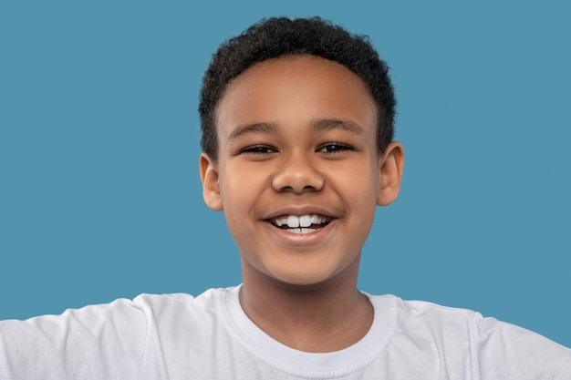 Gioia, umore. Gioioso ragazzo afroamericano con capelli ricci neri con sorriso a trentadue denti su sfondo blu