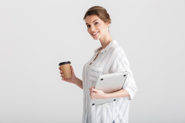 gioiosa bella donna che beve caffè da asporto e tiene in mano un laptop isolato su sfondo bianco