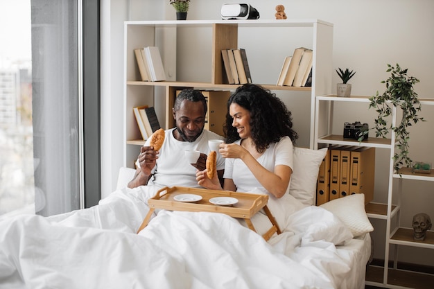 Gioiosa coppia sposata che mangia caffè e panini in camera da letto