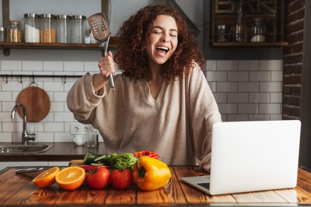 gioiosa donna caucasica che utilizza il computer portatile durante la cottura di insalata di verdure fresche nell'interno della cucina a casa