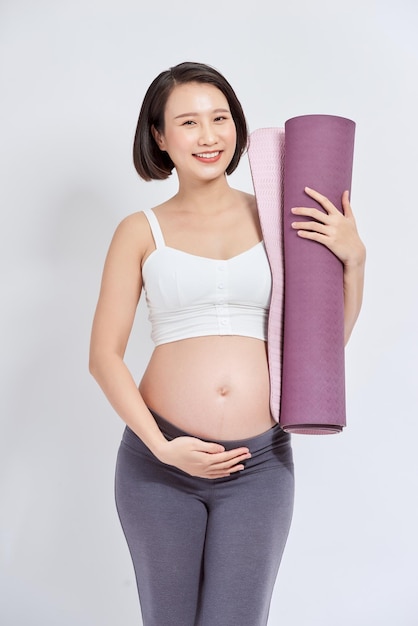 Gioiosa donna incinta azienda tappetino yoga sorridente alla telecamera in posa su sfondo bianco Stile di vita incinta e fitness