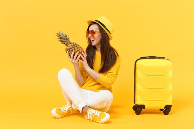 Gioiosa donna turistica viaggiatore in abiti estivi, cappello tenere frutta fresca matura ananas isolata su sfondo giallo arancio. Passeggero che viaggia all'estero durante il fine settimana. Concetto di viaggio in volo aereo.