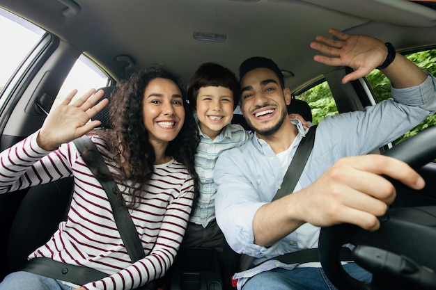 Gioiosa famiglia mediorientale con figlio piccolo che si diverte mentre guidano l'auto insieme, genitori arabi allegri e figlio maschio che agitano le mani alla telecamera, godendosi un viaggio su strada nel loro nuovo veicolo, primo piano