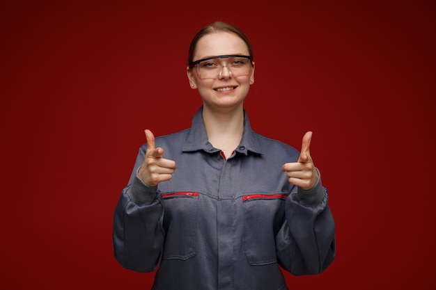 Gioiosa giovane bionda ingegnere femminile che indossa uniforme e occhiali di sicurezza che ti fanno gesto