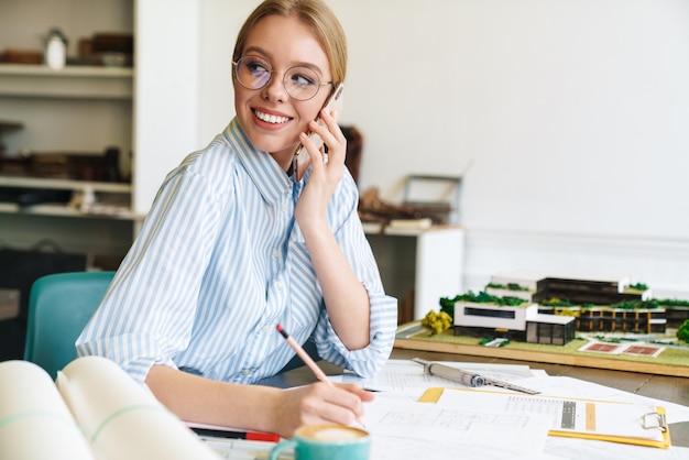 gioiosa giovane donna architetto in occhiali parlando al telefono cellulare durante la progettazione di bozze sul posto di lavoro