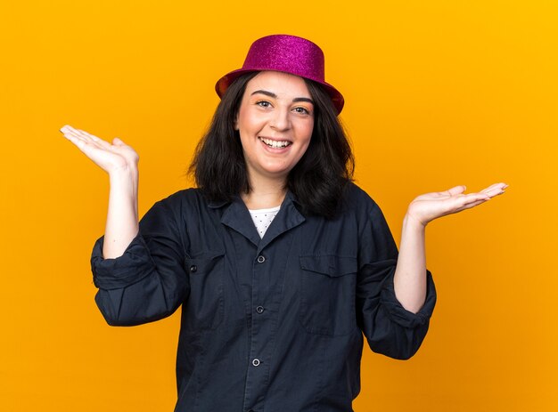 Gioiosa giovane donna caucasica da festa che indossa un cappello da festa guardando la parte anteriore che mostra le mani vuote isolate sulla parete arancione