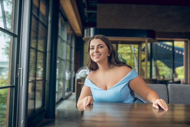 Gioiosa giovane donna dai capelli lunghi in abito blu in attesa di incontro al tavolo in caffè estivo