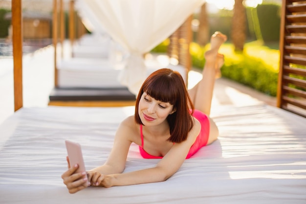 Gioiosa giovane donna in costume da bagno rosa sdraiata nel gazebo vicino alla piscina del resort e utilizzando lo smartphone