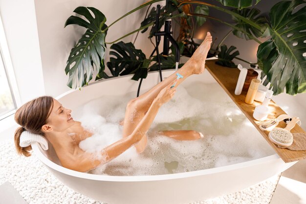Gioiosa giovane donna sdraiata nella vasca da bagno e che si rade le gambe con un rasoio da barba usa e getta per la cura del corpo