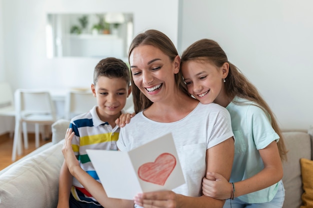 Gioiosa giovane madre e bambini piccoli seduti sul divano e abbracciati mentre leggono auguri e congratulazioni con le vacanze in una cartolina presentata a casa