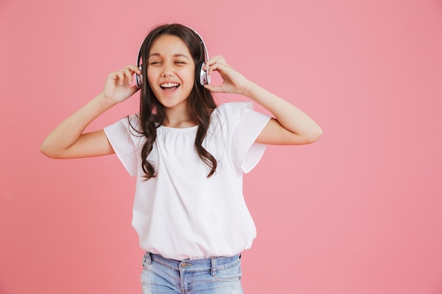 Gioiosa giovane ragazza 8-10 in abbigliamento casual cantando con gli occhi chiusi mentre si ascolta la musica tramite cuffie wireless, isolate su sfondo rosa