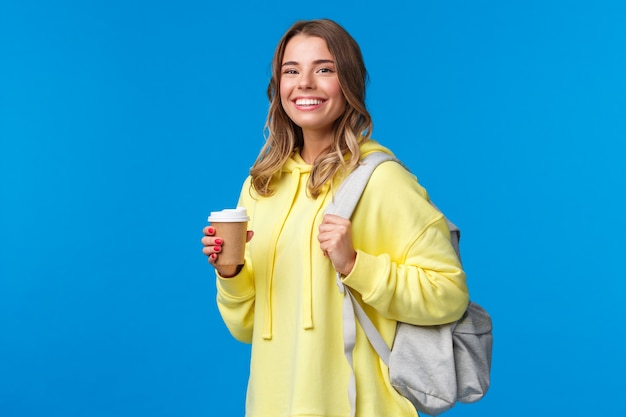 Gioiosa giovane studentessa bionda con zaino sorridente fotocamera soddisfatta come tenendo la tazza di caffè da asporto