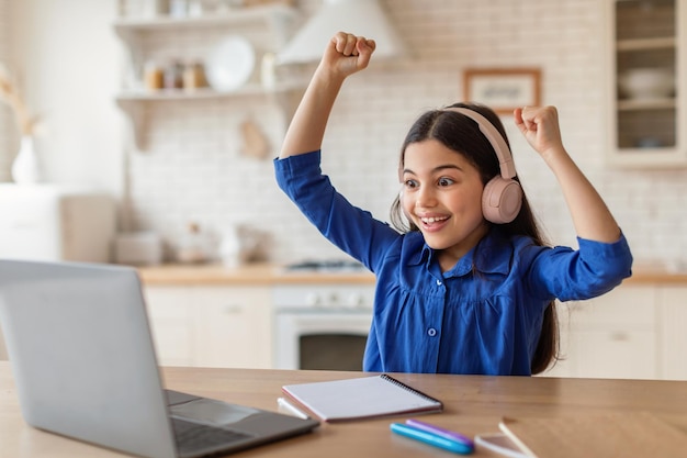 Gioiosa ragazza della scuola che impara online e celebra grandi notizie al coperto