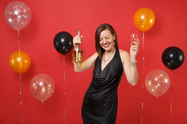 Gioiosa ragazza in abito nero che celebra tenendo il bicchiere, bottiglia di champagne su palloni ad aria di sfondo rosso brillante. Giornata internazionale della donna, felice anno nuovo, concetto di festa di compleanno mockup.