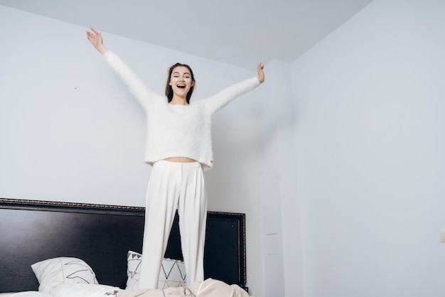 Gioiosa ragazza in pigiama bianco che salta sul letto la mattina presto sotto gli occhi macchie bianche