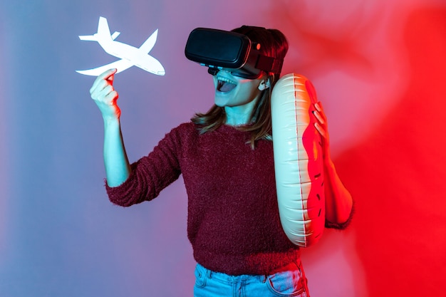 Gioiosa ragazza turistica con occhiali VR che tiene in mano un anello di gomma e un aeroplano simulato, viaggio virtuale