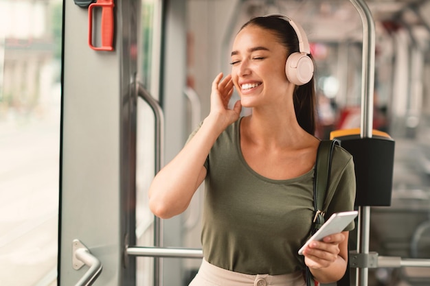 Gioiosa signora passeggero che tiene il cellulare che indossa le cuffie seduto in tram