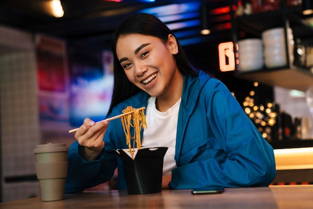 gioiosa simpatica donna asiatica che mangia noodles cinesi e sorride mentre è seduta al bar