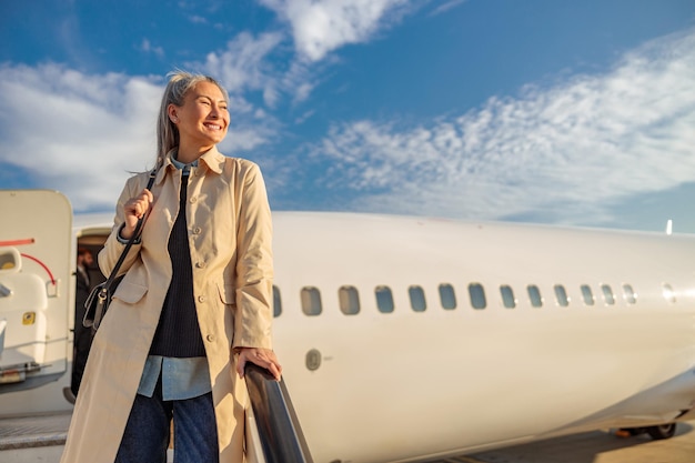 Gioiosa viaggiatrice che distoglie lo sguardo e sorride mentre si trova sulle scale d'imbarco dell'aeroplano sotto il cielo nuvoloso blu