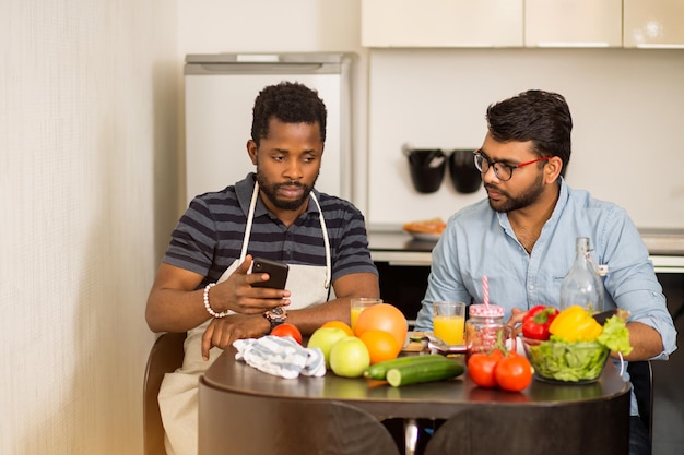 Gioiosi studenti multietnici che cucinano e si divertono in cucina. Giovane indiano e il suo amico afroamericano seduti a tavola, bevendo succo di frutta fresco, guardando la ricetta video, spettacolo culinario sul laptop.