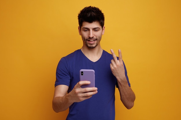 Gioioso giovane bell'uomo che prende selfie facendo gesto di pace su sfondo giallo