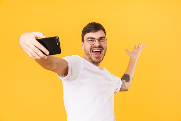 gioioso giovane in occhiali prendendo selfie sul cellulare e sorridente isolato su muro giallo