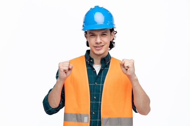 Gioioso giovane ingegnere maschio che indossa casco di sicurezza e giubbotto di sicurezza guardando la fotocamera facendo sì gesto isolato su sfondo bianco