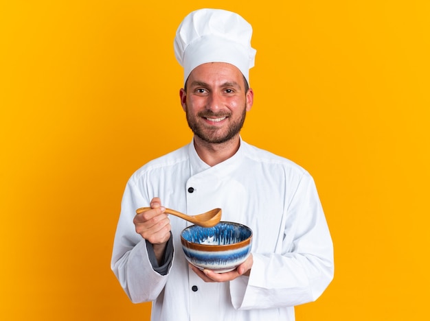 Gioioso giovane maschio caucasico cuoco in uniforme da chef e cappello che tiene ciotola e cucchiaio