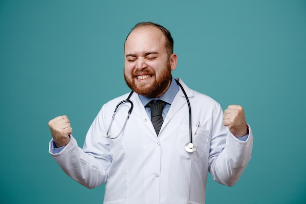 Gioioso giovane medico maschio che indossa un camice medico e uno stetoscopio intorno al collo che mostra un gesto di sì con gli occhi chiusi isolati su sfondo blu