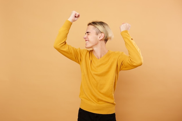 Gioioso giovane ragazzo biondo vestito con un maglione giallo è in posa in studio sullo sfondo beige.