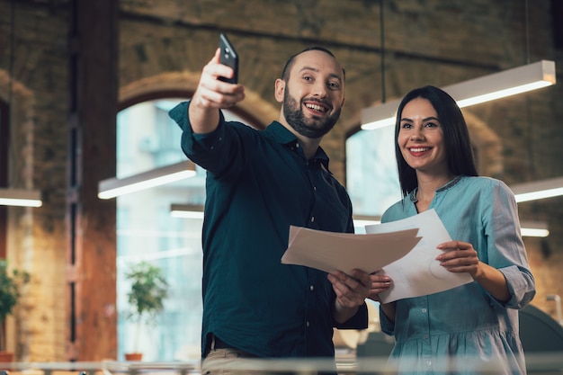 Gioioso giovane uomo e donna in possesso di fogli di carta e sorridente. Uomo con smartphone che punta in lontananza