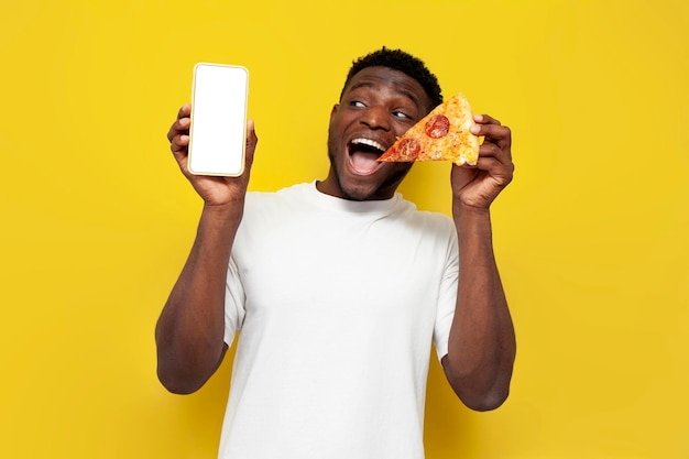 Gioioso uomo afroamericano che tiene la pizza e mostra lo schermo dello smartphone vuoto su sfondo giallo