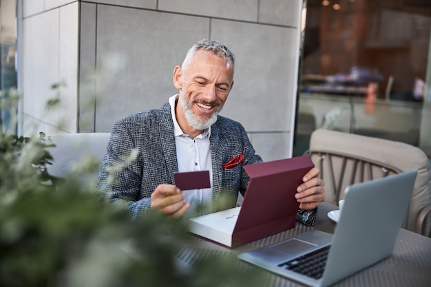 Gioioso uomo che invecchia guardando un cartellino rosso scuro in mano e sorride mentre è seduto in un caffè