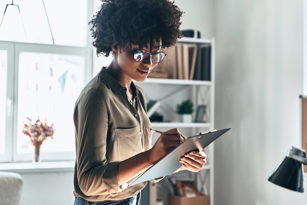 Giornata lavorativa impegnativa. Attraente giovane donna africana che scrive qualcosa mentre lavora in ufficio