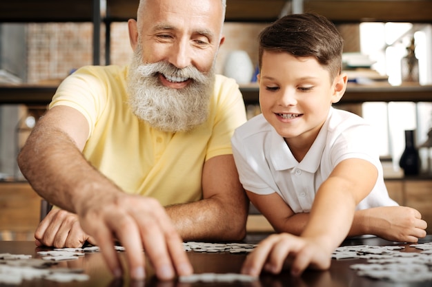 Giornata piacevole. Gioioso uomo anziano con la barba grigia seduto al tavolo accanto al nipote pre-adolescente e risolvendo un puzzle insieme a lui