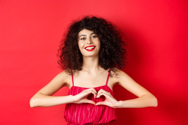 Giorno degli innamorati. Bella donna che celebra San Valentino, mostrando il segno del cuore e sorridente, in piedi in un romantico abito rosso su sfondo di studio.