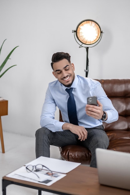 Giorno fortunato. Fiducioso giovane uomo d'affari sorridente guardando smartphone seduto in un ufficio moderno