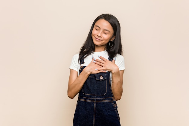 Giovane adolescente cinese carino Giovane donna bionda che indossa un cappotto contro una risata rosa mantenendo le mani sul cuore, concetto di felicità.