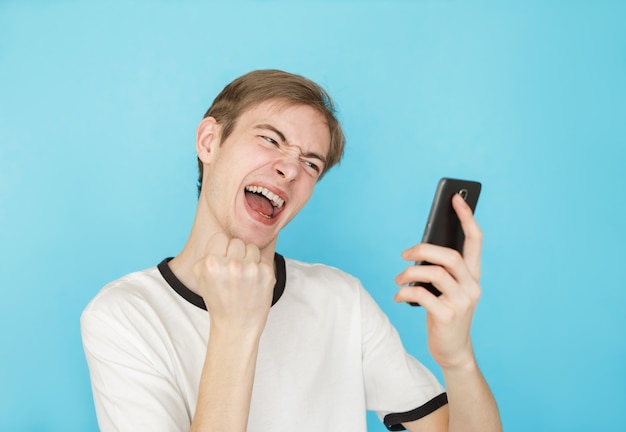 Giovane adolescente maschio divertente in maglietta bianca su sfondo blu guarda nello smartphone come se avesse vinto qualcosa
